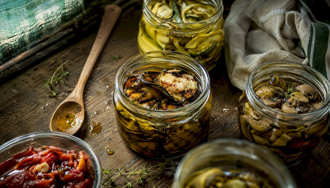 Geöffnete Gläser mit italienischen Antipasti in Öl stehen auf einer rustikalen Holzplatte.