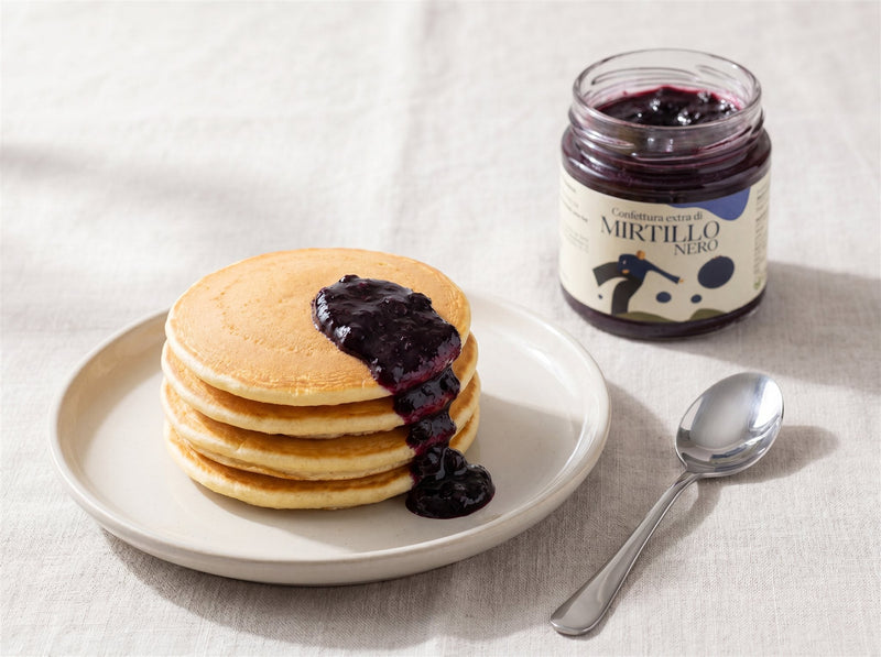 Pila di pancake soffici con confettura di mirtillo nero che cola sui lati e vasetto Minnelea aperto accanto