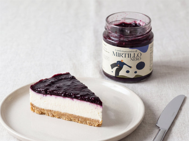 Fetta di cheesecake fredda con copertura di confettura di mirtillo nero su piatto chiaro, vasetto Minnelea sullo sfondo