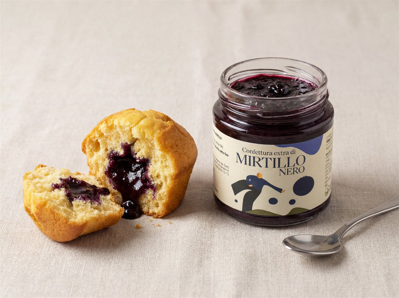 Muffin allo yogurt spezzato a metà che mostra il cuore di confettura di mirtillo nero, vasetto Minnelea accanto